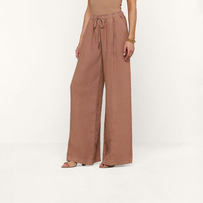 A person wearing the Connie Linen Drawstring Trouser—loose, wide-leg, brown linen pants featuring a drawstring waistband—paired with a sleeveless top and sandals, stands against a plain white background.