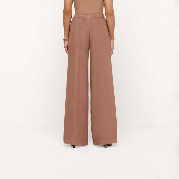 Standing against a white background, the person wears high-waisted, wide-leg Connie Linen Drawstring Trouser in brown paired with a sleeveless top. The view from the back highlights the full length of these vacation-ready pants.