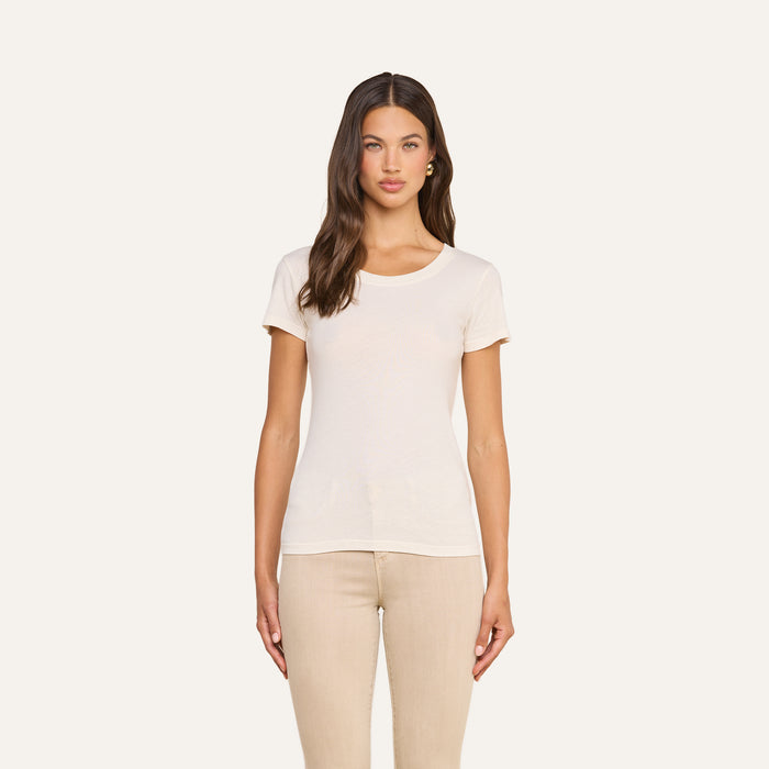 A woman with long brown hair stands facing forward against a plain light background, wearing the Cory Cotton Scoopneck Tee in vintage off-white—a short-sleeve, ultra-soft cotton classic—paired with beige pants.