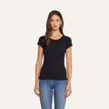A woman stands against a plain background, wearing the Cory Cotton Scoopneck Tee—crafted from ultra-soft black cotton—and blue jeans. She has long brown hair, a neutral expression, and faces forward with her arms relaxed at her sides.