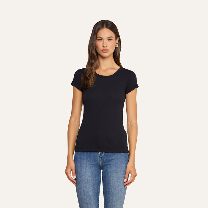 A woman stands against a plain background, wearing the Cory Cotton Scoopneck Tee—crafted from ultra-soft black cotton—and blue jeans. She has long brown hair, a neutral expression, and faces forward with her arms relaxed at her sides.