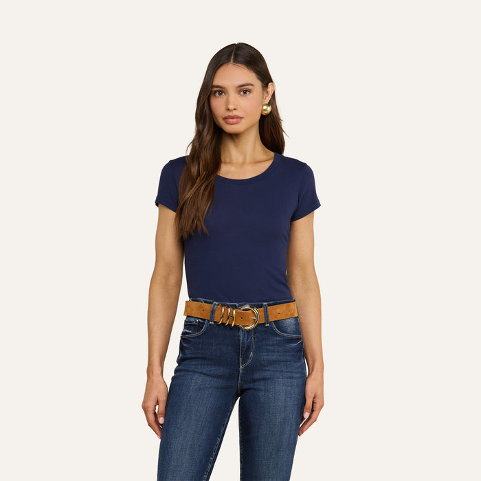 A woman with long brown hair is wearing a navy Cory Cotton Scoopneck Tee, blue jeans, a brown belt, and gold hoop earrings while standing against a plain off-white background.