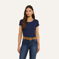 A woman with long brown hair is wearing a navy Cory Cotton Scoopneck Tee, blue jeans, a brown belt, and gold hoop earrings while standing against a plain off-white background.
