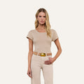 A woman with long blonde hair wears the Cory Cotton Scoopneck Tee in taupe, paired with light pants and a large gold-buckled belt, standing against a plain beige background with a neutral expression.