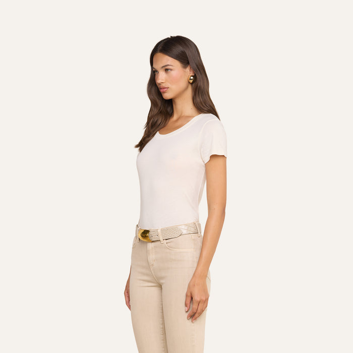 A woman with long brown hair stands in profile against a plain light background, wearing the ultra-soft Cory Cotton Scoopneck Tee in vintage off-white, paired with beige pants, gold earrings, and a beige belt with a gold buckle.