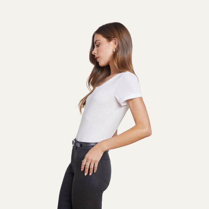 Wearing the Cory Cotton Scoopneck Tee, a woman stands in profile against a plain light background, her left hand tucked in the pocket of black pants and her long hair loose.