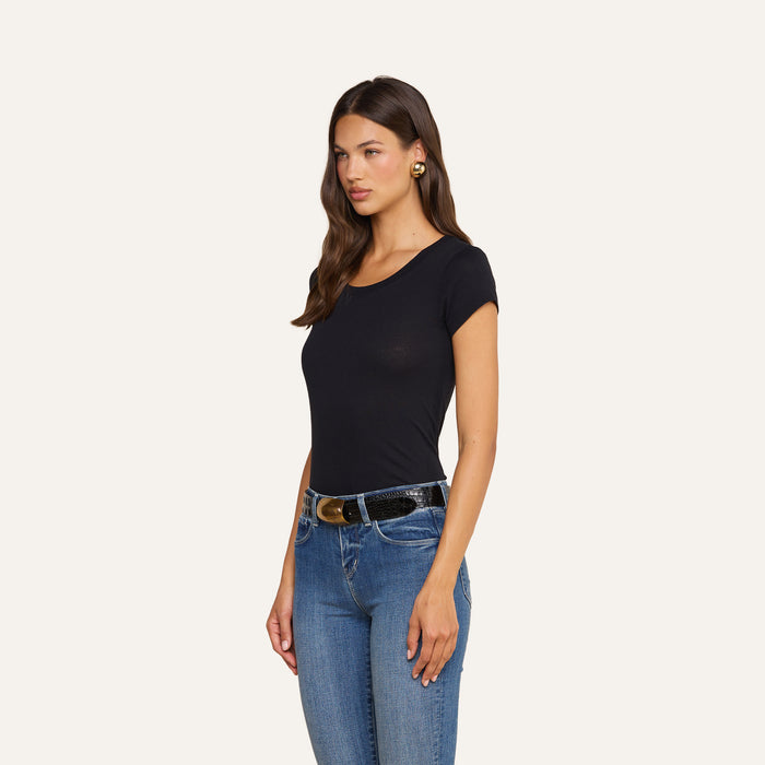 A woman with long brown hair wears the Cory Cotton Scoopneck Tee in black, paired with blue jeans and a black belt featuring a gold buckle. She stands against a plain light background, gazing slightly to the side.
