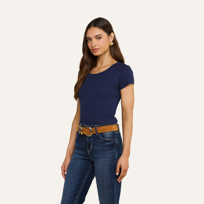 A woman with long brown hair and a neutral expression stands against a plain background, wearing the navy Cory Cotton Scoopneck Tee, paired with blue jeans, a brown belt, and gold earrings.