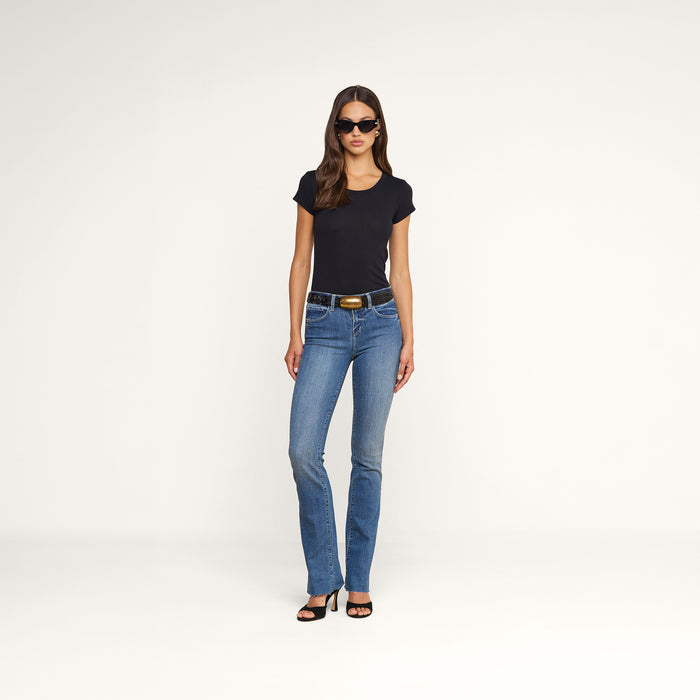 A woman stands against a plain white background, facing forward with her arms relaxed by her sides, wearing sunglasses, blue jeans, black heels, and the Cory Cotton Scoopneck Tee.