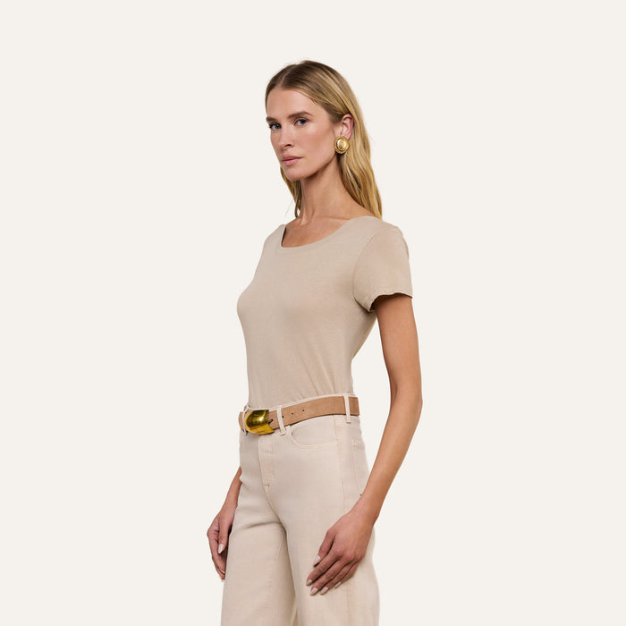 A woman with long blonde hair wears the Cory Cotton Scoopneck Tee in taupe, light beige pants, a tan belt with a gold buckle, and large gold earrings, standing against a plain beige background.