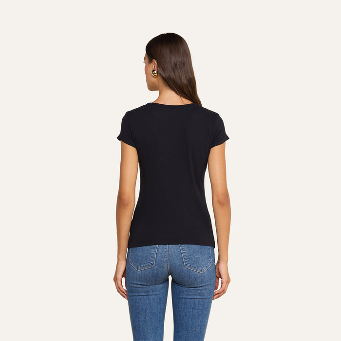 A woman with long brown hair, wearing the Cory Cotton Scoopneck Tee in black and blue jeans, stands facing away from the camera against a plain background.
