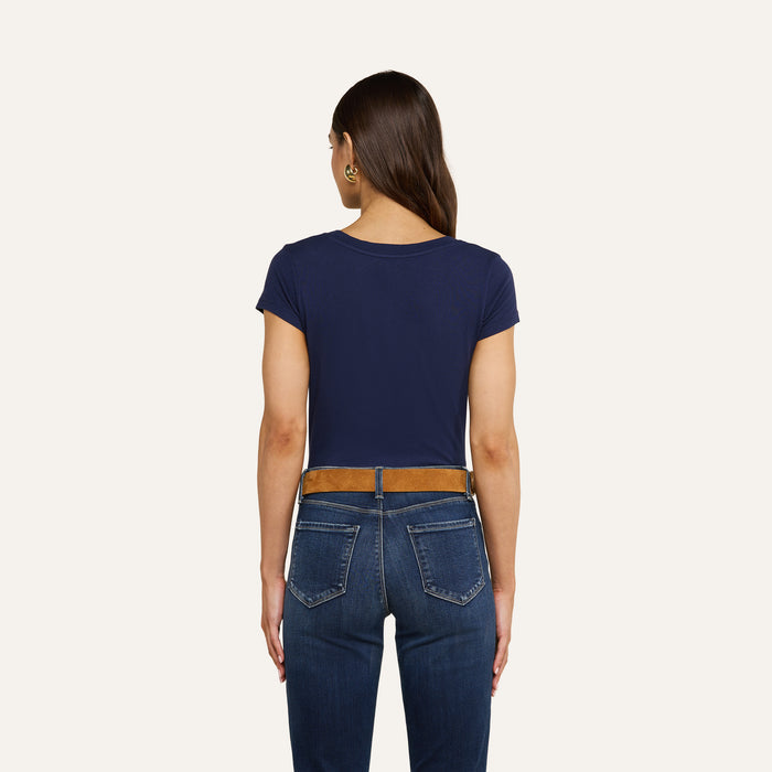 A woman with long brown hair, wearing the Cory Cotton Scoopneck Tee in navy, paired with blue jeans and a brown belt, stands facing away from the camera against a plain light background.