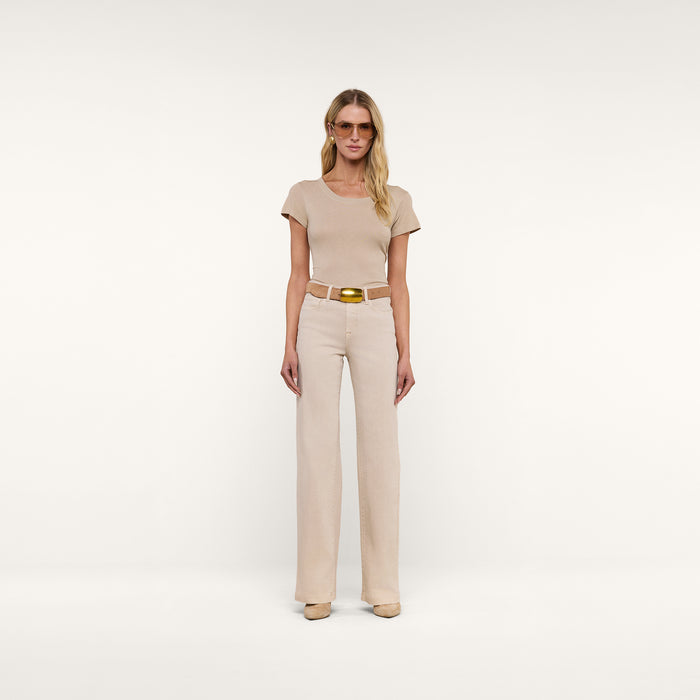 A woman with long blonde hair and a relaxed expression stands against a beige background wearing the Cory Cotton Scoopneck Tee in taupe, high-waisted light beige pants with a brown belt, beige shoes, sunglasses, and earrings.