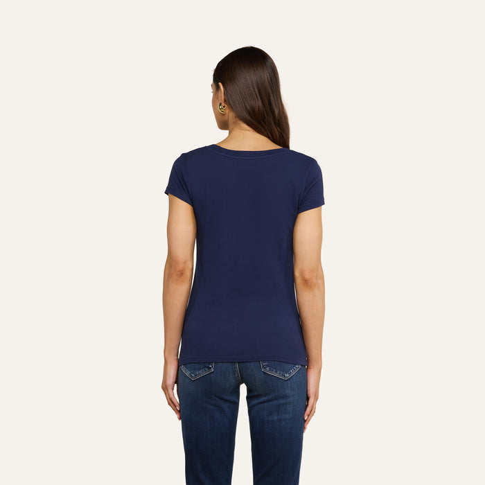 A woman with long brown hair is standing with her back to the camera, wearing a navy Cory Cotton Scoopneck Tee and blue jeans against a plain white background.