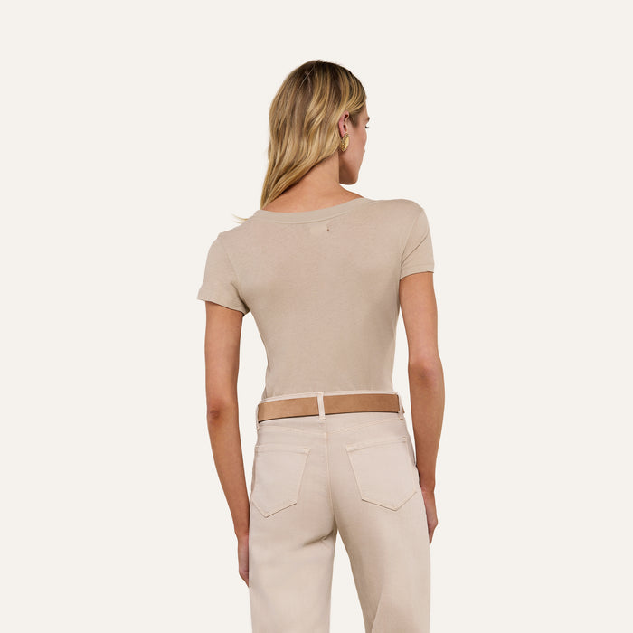A woman with blonde hair stands facing away, wearing the Cory Cotton Scoopneck Tee in taupe and high-waisted light jeans with a brown belt, set against a neutral background.