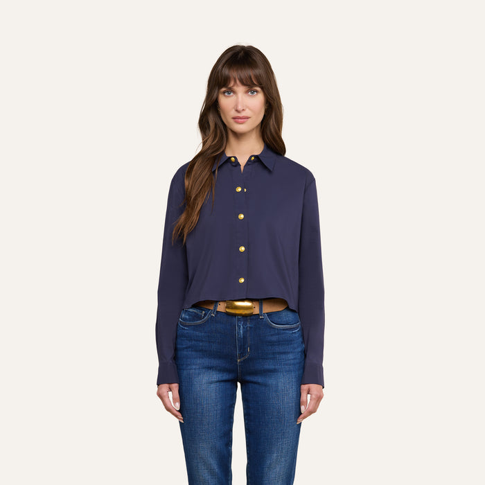 A woman with long brown hair stands facing forward against a plain light background, wearing a navy blue Cosette Shirt and blue jeans, her expression neutral.
