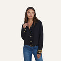 Wearing the Cosette Shirt—a tailored black button-up—a woman with long brown hair stands against a plain background, looking to her left. She pairs the top with blue jeans and chunky gold bracelets.