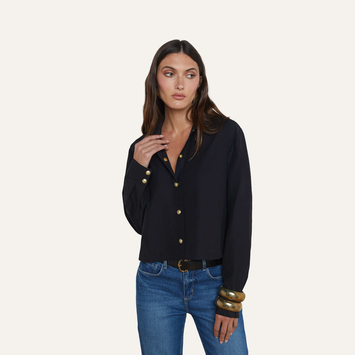 Wearing the Cosette Shirt—a tailored black button-up—a woman with long brown hair stands against a plain background, looking to her left. She pairs the top with blue jeans and chunky gold bracelets.
