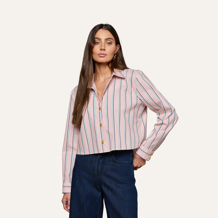 A woman with long brown hair is wearing the Cosette Shirt—a pink tailored shirt with vertical blue and red stripes, featuring a cropped high-low hem—paired with dark blue jeans and standing against a plain light background.