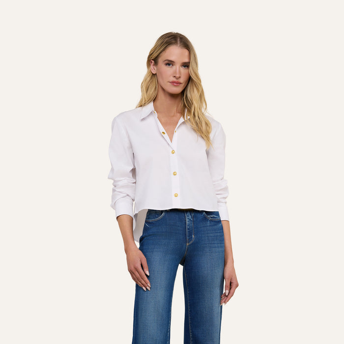 A woman with long blonde hair wears the Cosette Shirt, a tailored white button-up, paired with blue jeans as she stands against a plain, light background.