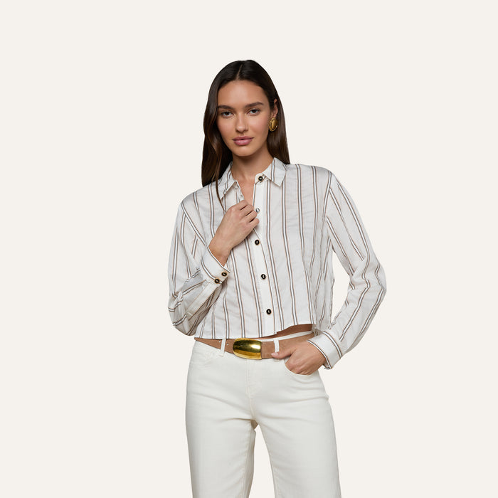 A woman wears the Cosette Shirt with jacquard stripes, paired with white pants, standing against a plain background—one hand near her collar and the other relaxed by her side.