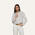 A woman wears the Cosette Shirt with jacquard stripes, paired with white pants, standing against a plain background—one hand near her collar and the other relaxed by her side.