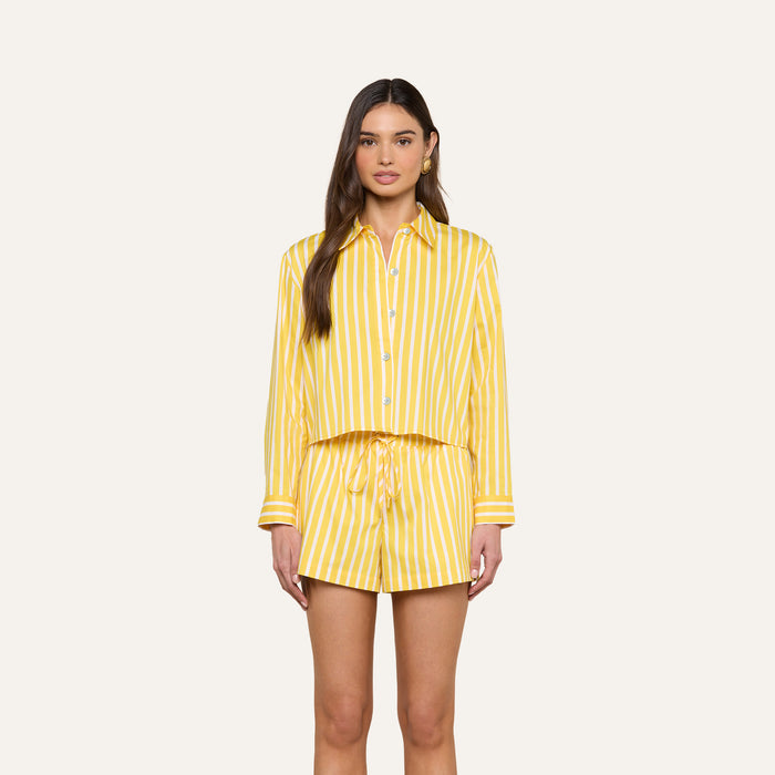 A woman stands facing forward, wearing the Cosette Shirt—a matching yellow and white vertical striped organic cotton button-up—paired with drawstring shorts against a plain light background.