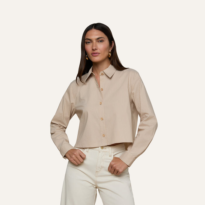 A woman stands with her hands on her hips, wearing the Cosette Shirt—a tailored, beige long-sleeve button-up—paired with off-white pants. She has long, straight brown hair and faces forward against a plain light background.