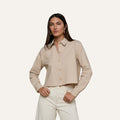 A woman stands with her hands on her hips, wearing the Cosette Shirt—a tailored, beige long-sleeve button-up—paired with off-white pants. She has long, straight brown hair and faces forward against a plain light background.