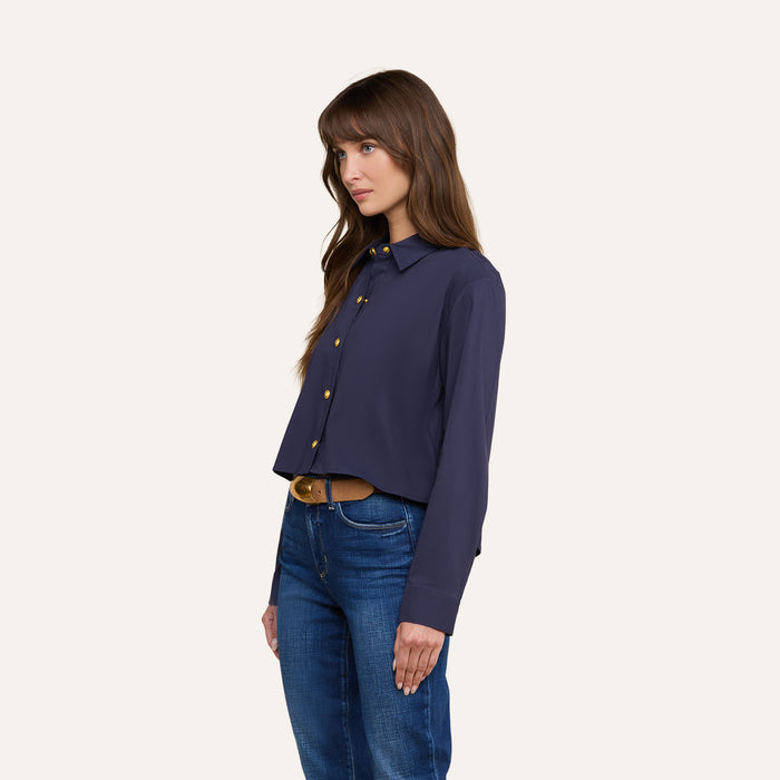 A woman with long brown hair wears the Cosette Shirt in navy, paired with blue jeans, standing against a plain light background and facing slightly left with a neutral expression.
