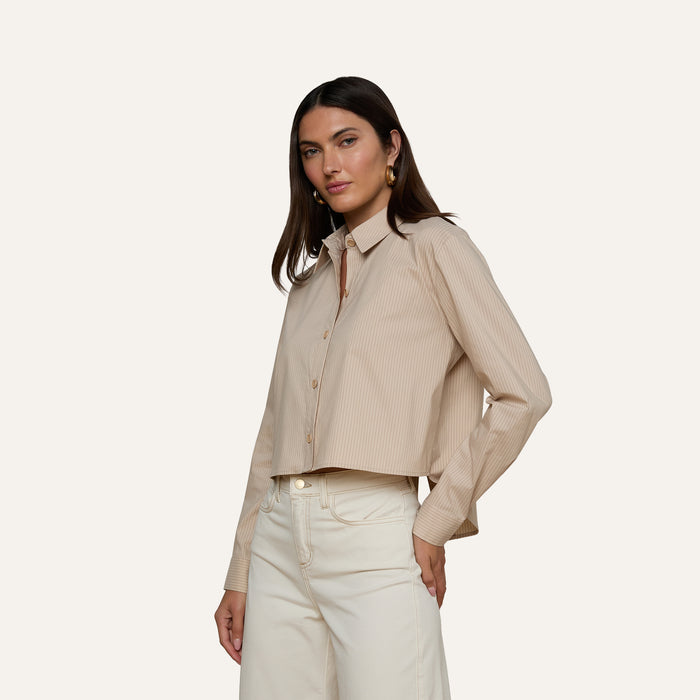 Wearing the Cosette Shirt, a beige, long-sleeve organic cotton top with subtle stripes, a woman pairs it with high-waisted cream pants. She stands against a plain light background, looking confidently at the camera.