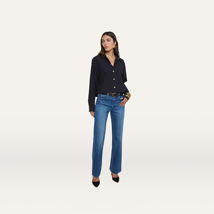 A woman stands against a plain white background wearing the Cosette Shirt, styled with blue jeans, a black belt, black heels, and large hoop earrings.