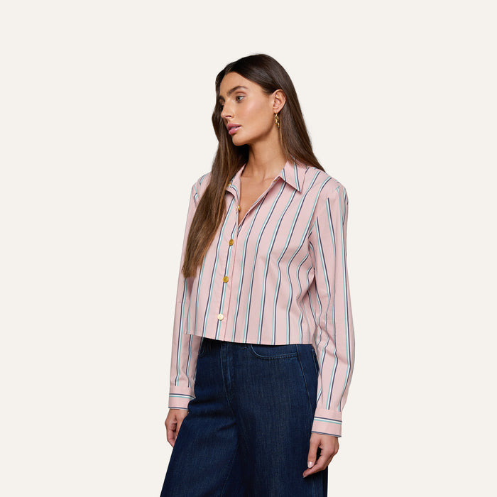 A woman wears the Cosette Shirt, a pink long-sleeve, button-up tailored shirt with vertical stripes, paired with dark wide-leg jeans as she stands against a plain light background.