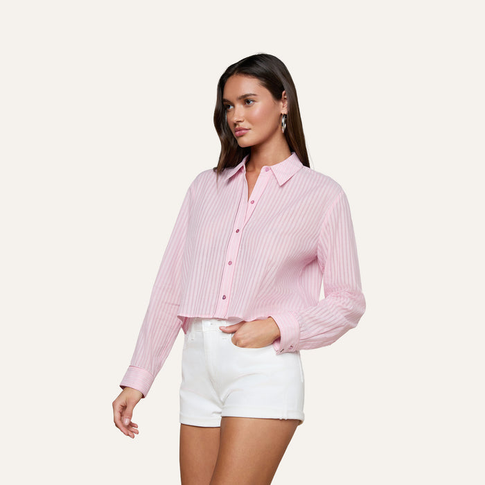 A woman wearing the Cosette Shirt—a light pink, long-sleeve shirt with white vertical stripes—paired with white high-waisted shorts, poses against a plain off-white background.