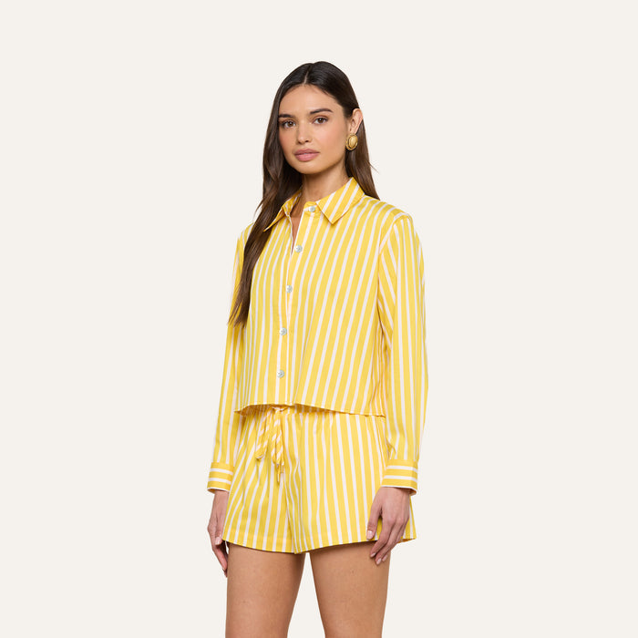 A woman wearing the Cosette Shirt—yellow and white vertically striped and tailored—with matching striped drawstring shorts, standing against a plain light background.