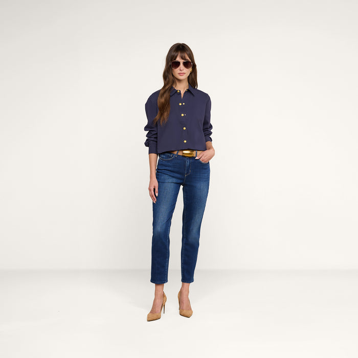 A woman stands against a plain white background, hands in pockets and looking forward, wearing sunglasses, blue jeans, beige heels, and the Cosette Shirt in navy.