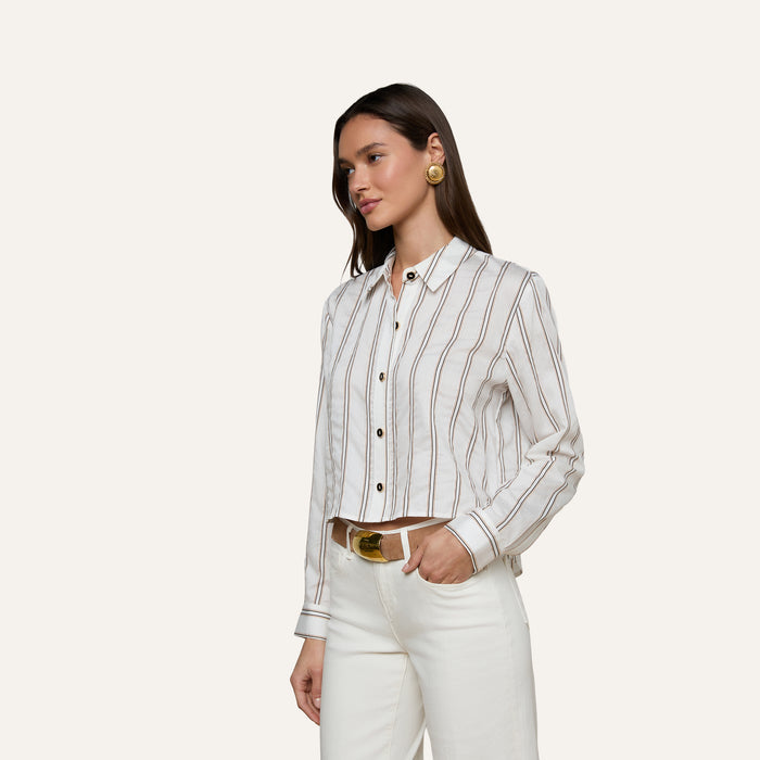 A woman wears the Cosette Shirt—a white button-up tailored shirt with brown jacquard stripes—paired with white pants, a tan belt, and large round earrings, standing against a plain light background.