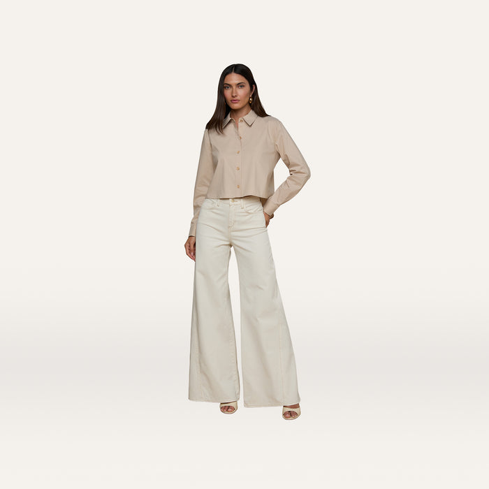 A woman stands against a plain white background, wearing the Cosette Shirt made from organic cotton with long sleeves, paired with high-waisted, wide-leg white pants. She has long dark hair and is wearing open-toed heels.