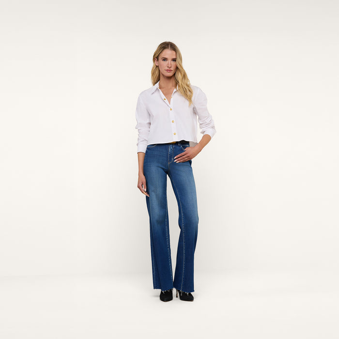 A woman with long blonde hair stands in a relaxed, casual pose against a plain white background, wearing the Cosette Shirt—a tailored white button-down—paired with blue high-waisted jeans and black shoes.