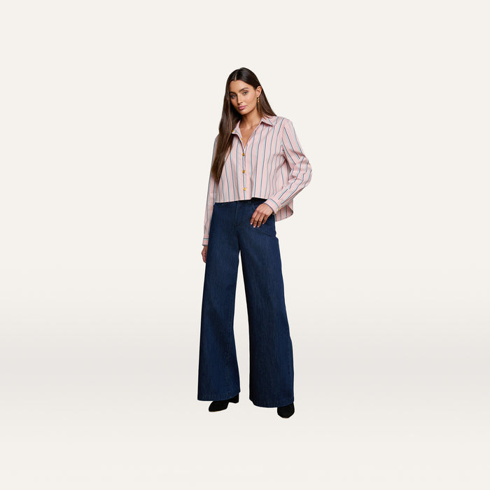 A woman stands against a plain white background wearing the Cosette Shirt—a tailored, striped button-up made from an organic cotton-blend—paired with high-waisted, wide-leg dark blue jeans and black shoes.
