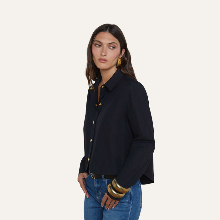 A woman with long brown hair wears the black Cosette Shirt, a stretch cotton-blend tailored top, paired with blue jeans and accented by large gold bracelets and gold earrings as she stands against a plain light background.