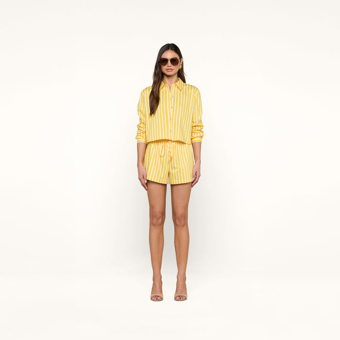 A woman wearing sunglasses and the Cosette Shirt, a yellow and white vertical striped tailored shirt with sleeves rolled up, paired with matching shorts and nude high-heeled sandals, stands against a plain white background.
