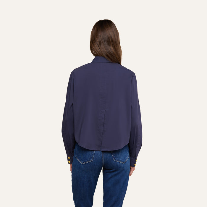 A woman with long brown hair is shown from the back, wearing the Cosette Shirt—a dark blue tailored shirt with gold buttons on the cuffs—paired with blue jeans, standing against a plain light background.