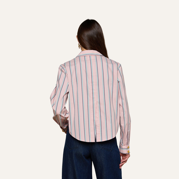 A woman with long brown hair is seen from behind, wearing the Cosette Shirt—a tailored pink, long-sleeve, collared shirt featuring vertical blue and white stripes and a cropped high-low hem—paired with dark blue wide-leg pants against a plain light background.