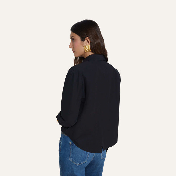 A woman with long brown hair wears the Cosette Shirt—a black stretch cotton-blend tailored design—paired with blue jeans and large gold earrings, standing facing away with her head turned slightly to the side against a plain light background.