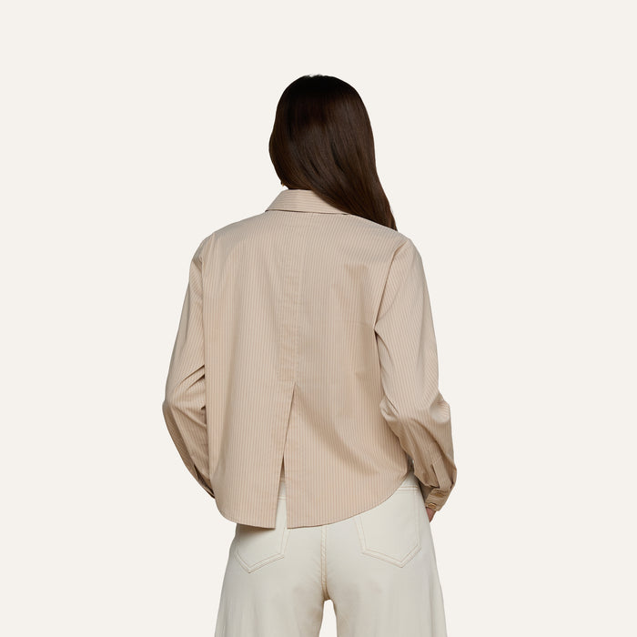 A person with long brown hair, seen from the back, wears the Cosette Shirt—a beige, collared, tailored shirt with long sleeves and a back slit—paired with white pants. The background is plain and light-colored.