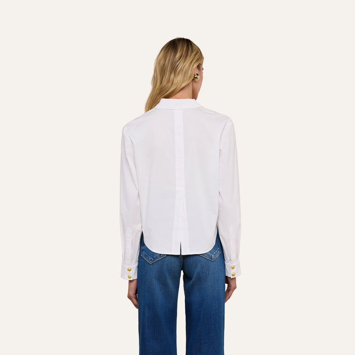 A person with long blonde hair, seen from behind, wears the Cosette Shirt—a white organic cotton tailored shirt featuring gold cuff buttons and a center split at the hem—paired with blue jeans. The background is plain and light-colored.