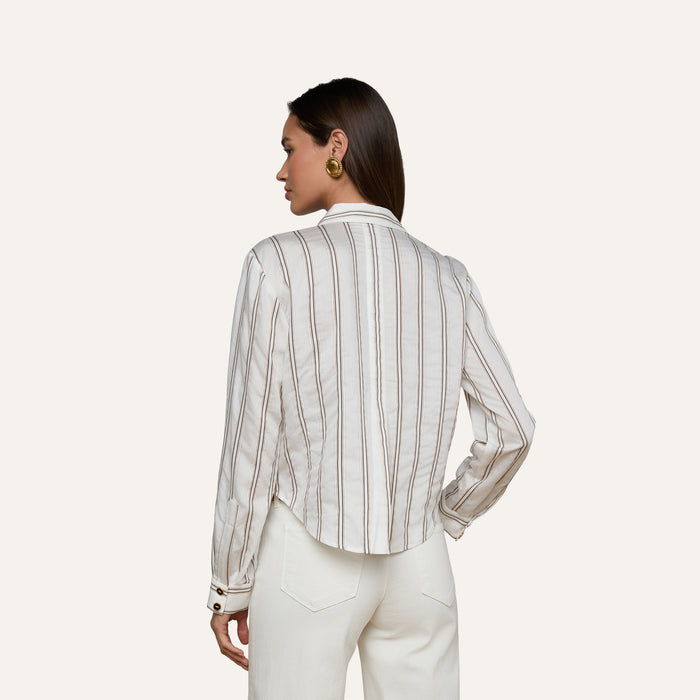 A woman stands facing away, wearing the Cosette Shirt—a tailored white long-sleeve top with vertical jacquard stripes—paired with white pants. She has straight brown hair and accessorizes with gold round earrings, set against a plain, light-colored background.