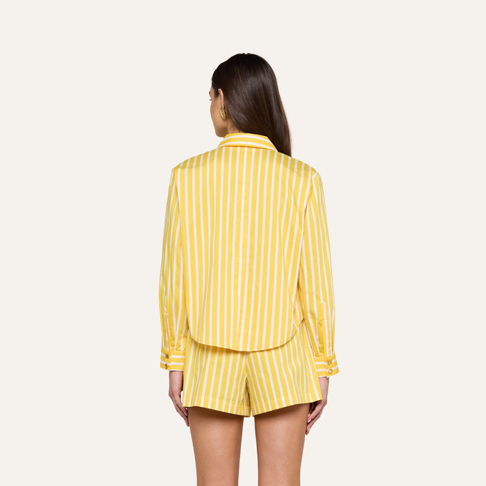 A woman stands facing away, wearing the yellow and white vertical striped Cosette Shirt with matching shorts against a plain, light-colored background.