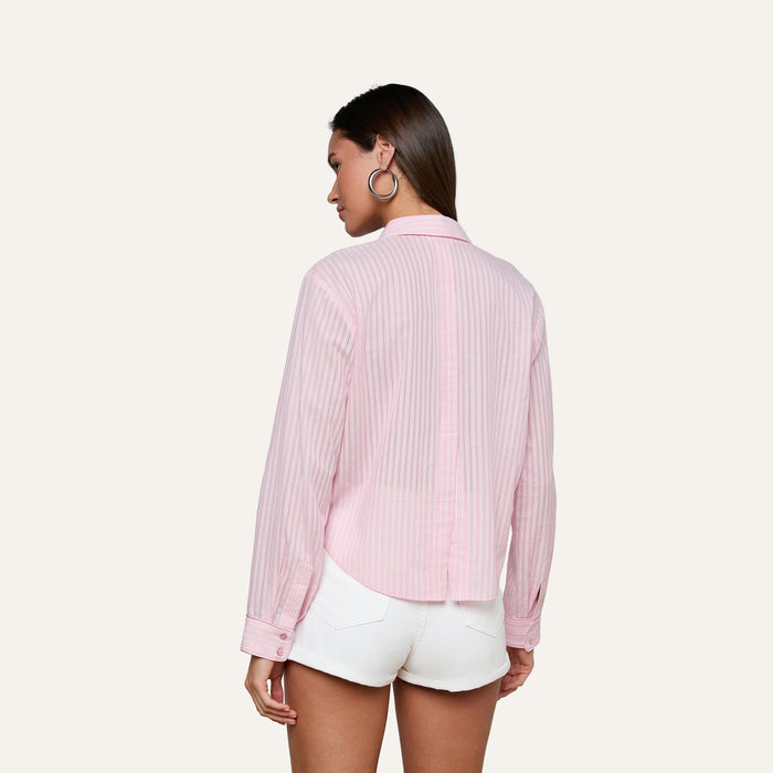 A woman stands facing away, wearing the Cosette Shirt in light pink stripes paired with white shorts. She has shoulder-length hair and large hoop earrings against a plain light background.